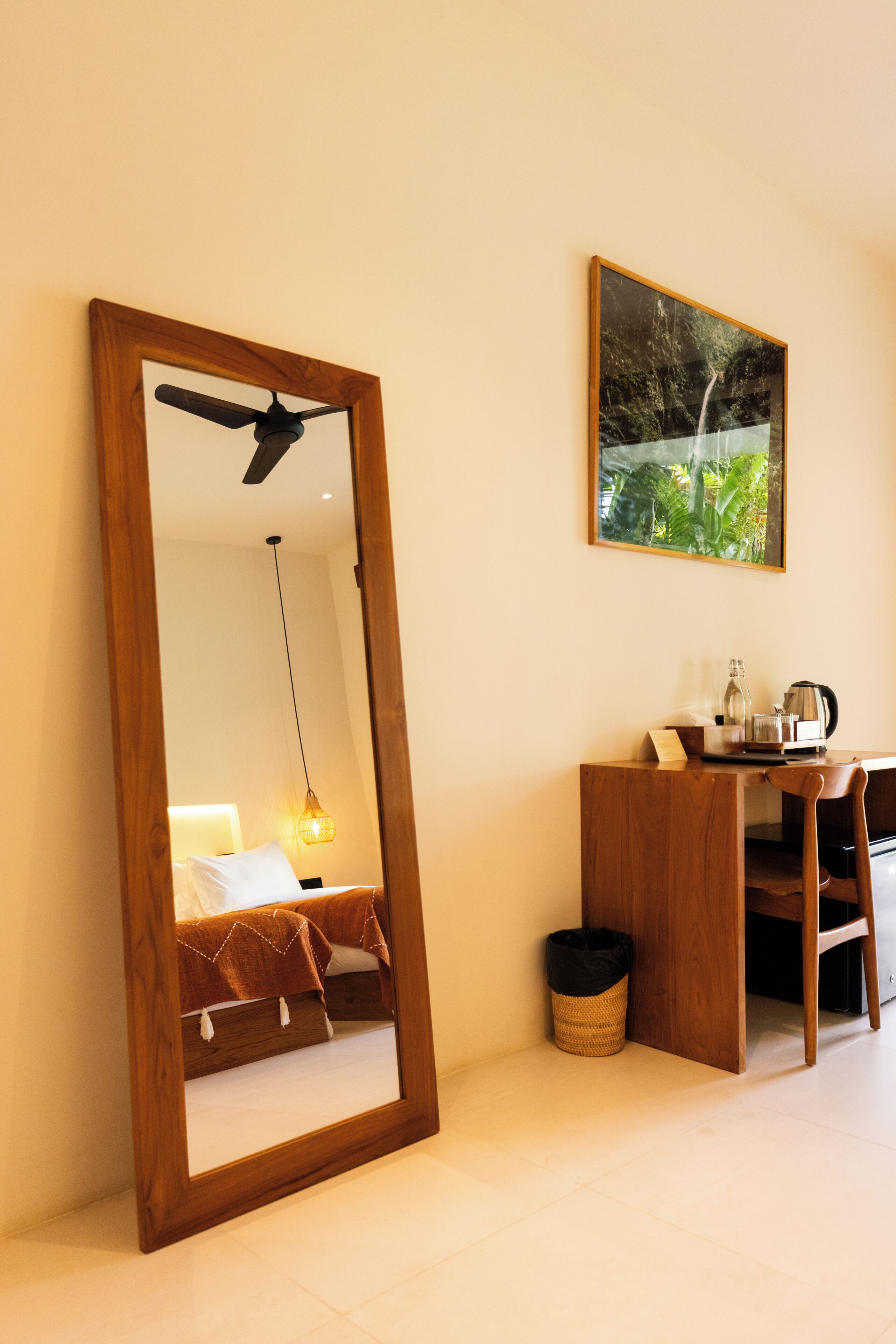 A room with a large mirror, a bed with a headboard, a desk with a chair, a hanging light, and a framed picture on the wall.