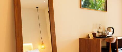 A room with a large mirror, a bed with a headboard, a desk with a chair, a hanging light, and a framed picture on the wall.