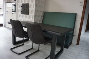 Un comedor moderno con una mesa y dos sillas, un banco acolchado verde, y una cocina al fondo.