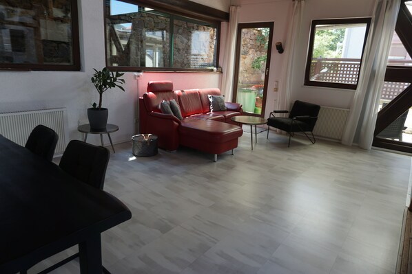 Amplia sala de estar con un sofá rojo, un sillón negro y una pequeña mesa redonda. Hay una planta en maceta sobre una mesita auxiliar y una ventana con vistas al exterior.