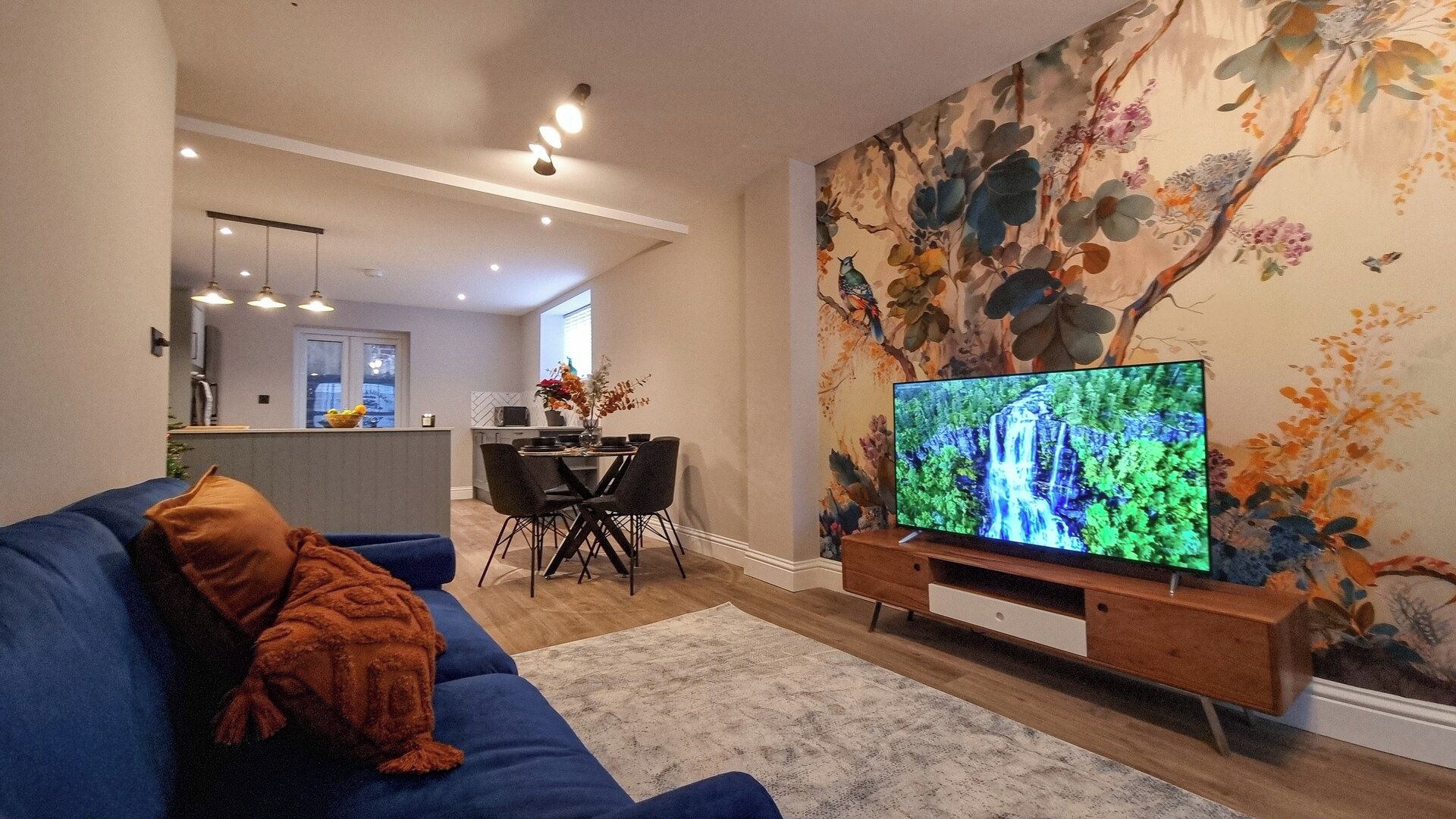 Una sala de estar con un sofá azul, un televisor sobre un soporte de madera y un mural floral.