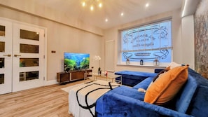 A modern living room with a flat-screen TV, a blue sofa, and a large window with blinds. - Heart of Brighton: 4 Bed, 4 Bath + Parking (Brighton and Hove)