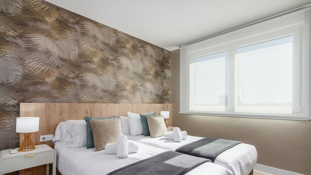 Habitación de hotel con dos camas, cabecero de madera, mesita de noche con lámpara y un ventanal grande.