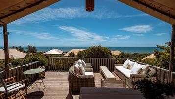 Eine Holzterrasse mit Gartenmöbeln, darunter eine Sitzecke, Stühle und ein Tisch, mit Blick auf einen Strand und den Ozean.