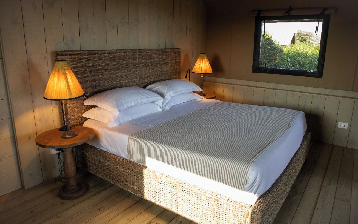 Una cama individual con estructura de mimbre, dos lámparas de noche y una ventana con vistas a la vegetación.