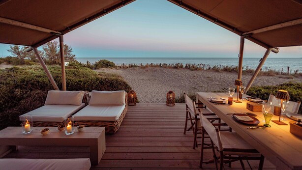 A tented area with a wooden deck, two lounge chairs, a dining table set for two, and a view of the beach and ocean. - Resort & Glamping Terre di Sacra (Capalbio)