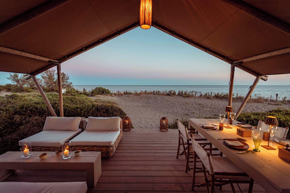 Un área con carpas, una terraza de madera, dos sillones, una mesa de comedor para dos personas y vistas a la playa y al océano.