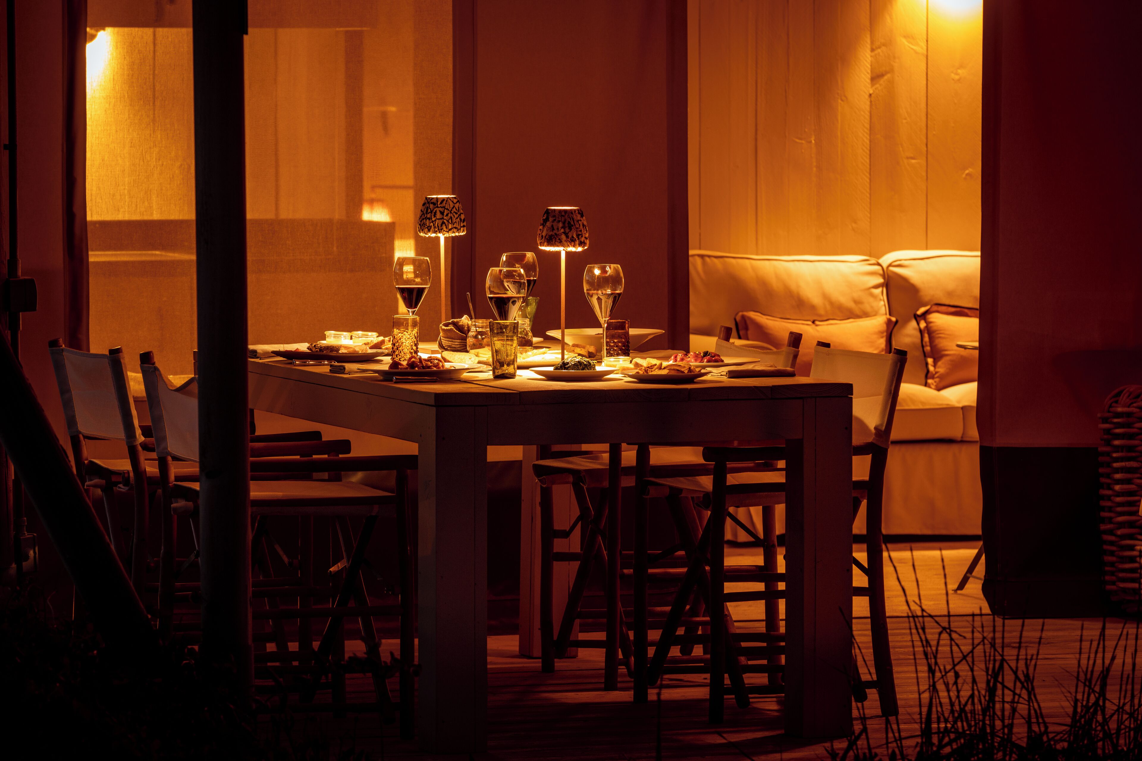 A dining area with a table set for six, surrounded by chairs, with a sofa and a lamp in the background.