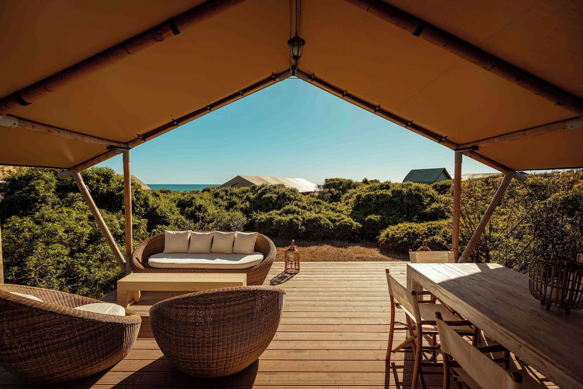 Un área de estar al aire libre con una mesa de madera y sillas de mimbre, un sofá acolchado y un farol en una terraza de madera con vistas a una zona boscosa.