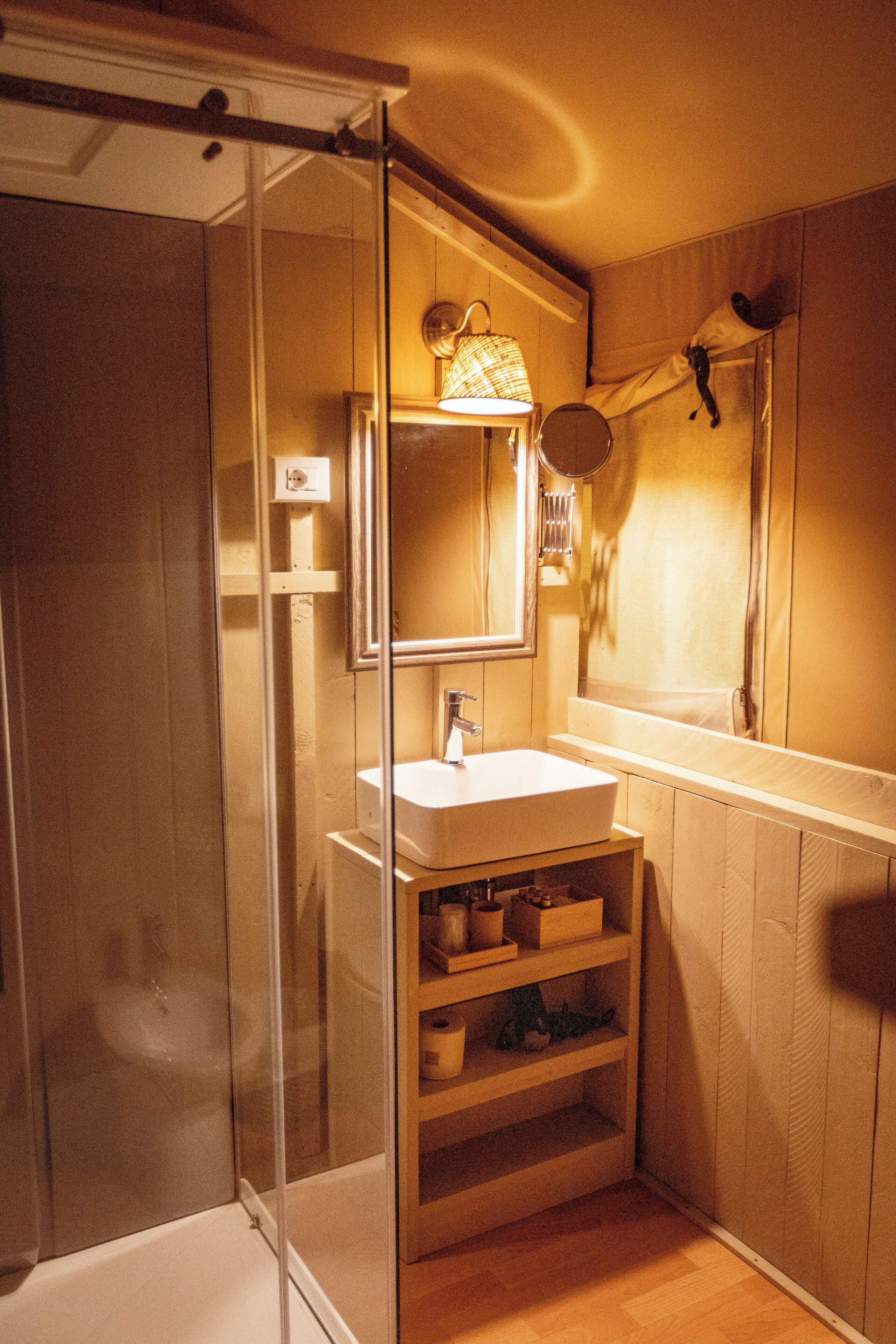 Un baño con cabina de ducha de vidrio, un lavabo blanco, un espejo y estantes de madera.
