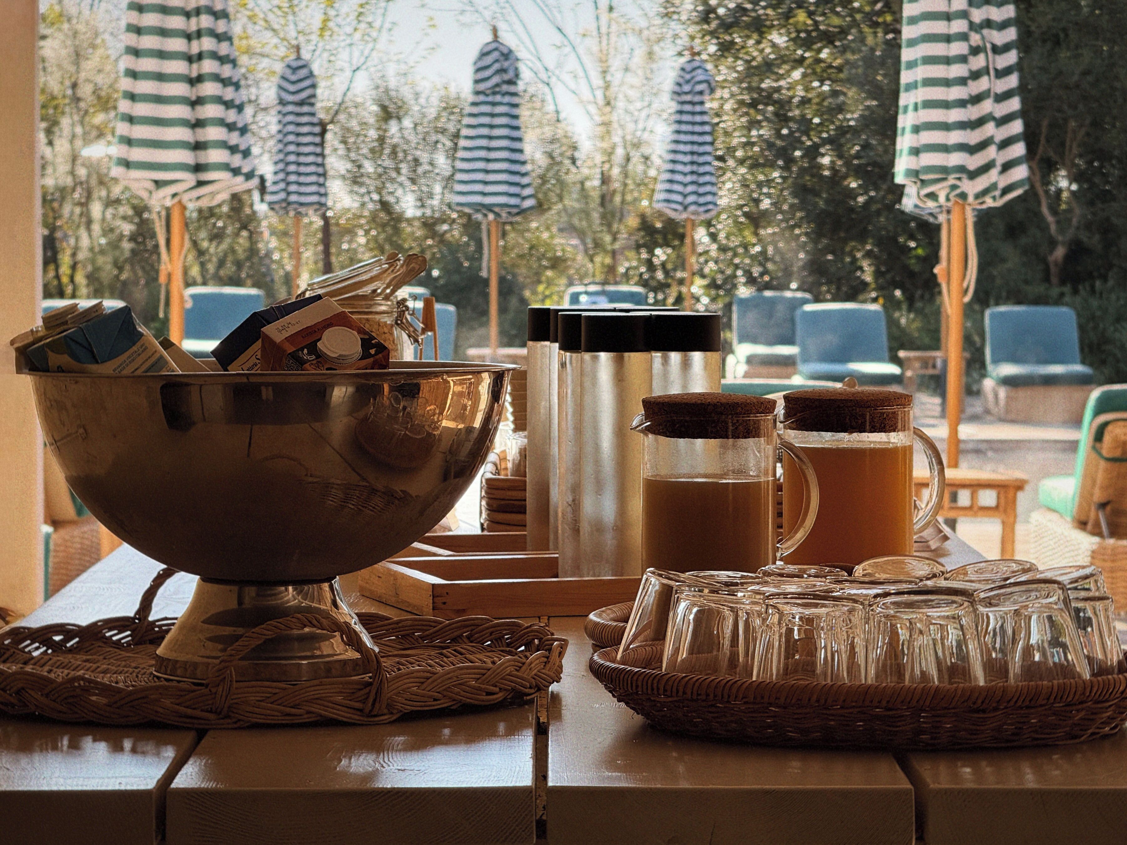 Uma mesa com uma tigela de metal, uma cafeteira e um jarro de vidro, rodeada por cestos de vime e cadeiras azuis sob toldos listrados.