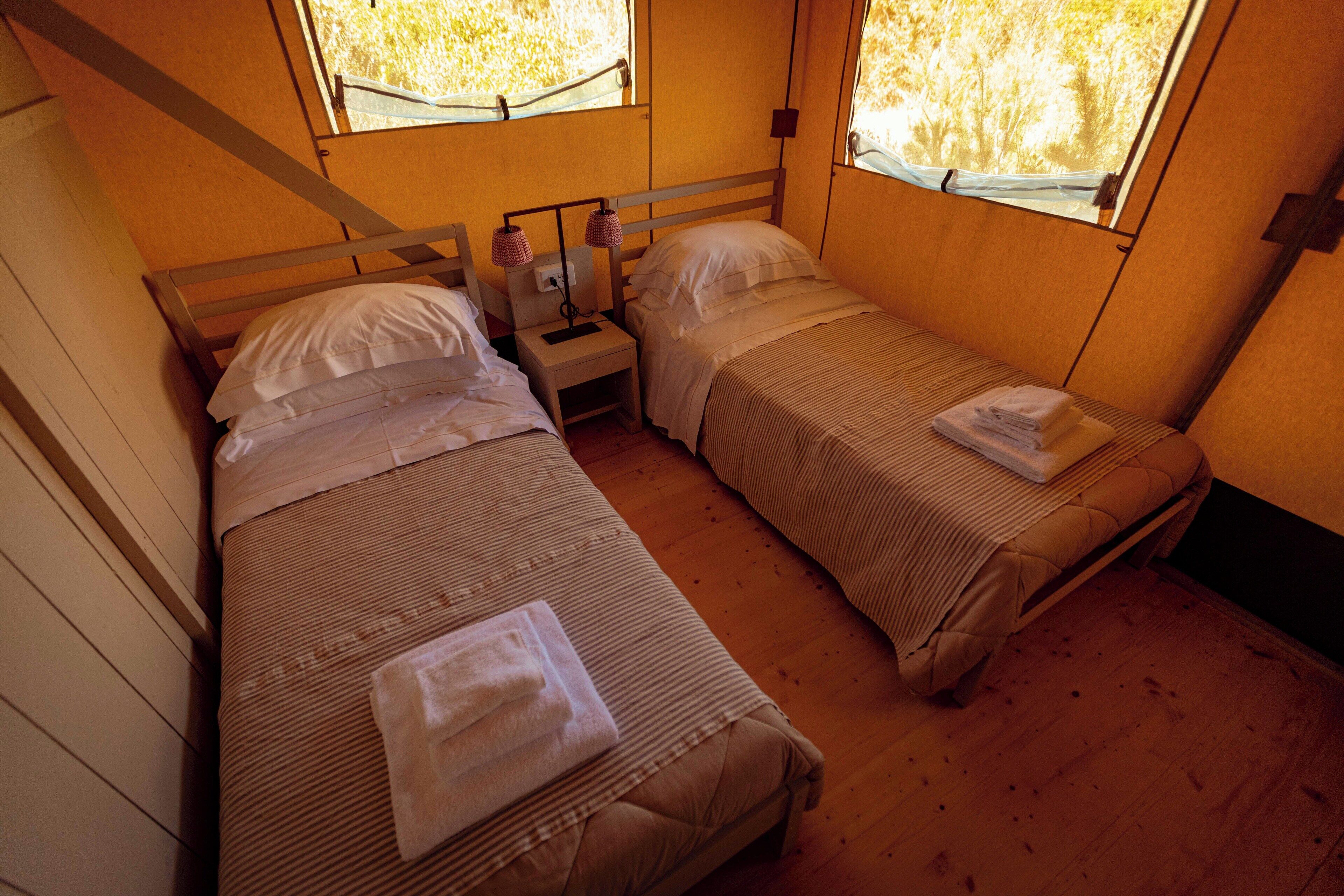 A room with two beds, each with a pillow and a folded towel. There is a nightstand with a lamp between the beds.