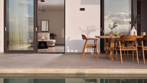 A modern outdoor dining area with a pool, wooden furniture, and a glass door leading to a living room.