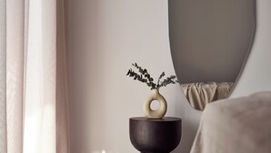 A modern bedroom with a large mirror, a bedside table with a vase of greenery, and a bed with a beige blanket.