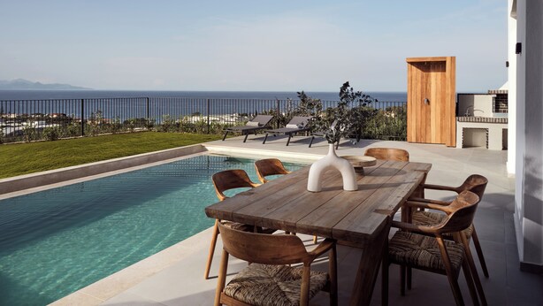 A poolside area with a wooden table and chairs, a vase, and a view of the sea.