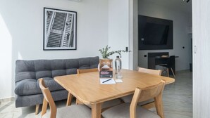 Een moderne woonkamer met een grijze bank, een houten eettafel met vier stoelen, een lijst met een afbeelding aan de muur, en een televisie op de achtergrond.