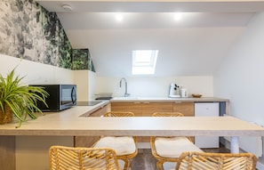 Eine moderne Küche mit einer zentralen Kochinsel, einer Mikrowelle, einem Spülbecken und einem kleinen Kühlschrank. Um die Insel stehen Korbstühle und auf der Arbeitsplatte steht ein Blumentopf.