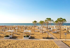 Une plage avec des chaises longues et des tables blanches disposées en rangées, une promenade en bois et quelques arbres.