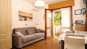 A modern living room with a sofa, a dining table with chairs, a kitchen area with a microwave, and a sliding glass door leading to an outdoor area. - Stylish Lagoonside studio retreat in Lignano (Lignano Sabbiadoro)