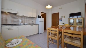 Eine kompakte Küche mit weißen Schränken, einer Mikrowelle und einem Kühlschrank. Es gibt einen Esstisch mit Stühlen und ein farbenfrohes Sofa mit einem Hut darauf.