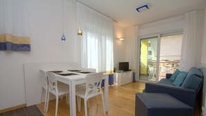 A modern living room with a sofa, a dining table with chairs, a TV, and a balcony with a view. - Modern apartment with solarium near the beach (Bibione)