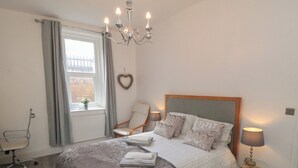 Une chambre à coucher avec un lit, une chaise, une table, un lustre et une fenêtre avec des rideaux.