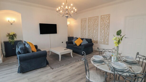 Un salon moderne avec un lustre, deux canapés, une table basse, un téléviseur, des panneaux décoratifs et un coin repas avec des chaises et de l’assiette.
