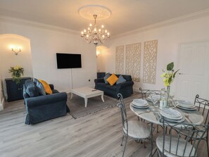A modern living room with a chandelier, two sofas, a coffee table, a TV, decorative panels, and a dining area with chairs and plates.
