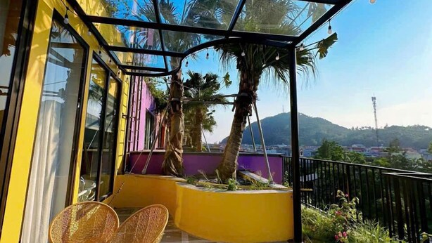 Ein sonnendurchfluteter Balkon mit Glasveranda, ausgestattet mit zwei Korbstühlen, einem kleinen Tisch und Blick auf Palmen und ferne Hügel.