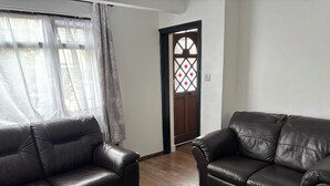 A living room with two dark leather sofas, wooden flooring, a door with a decorative glass panel, and sheer curtains. - Dagenham - Stay 5 - Garden - Parking - Petfriendly (Dagenham)