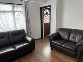 Un salón con dos sofás de cuero oscuro, suelo de madera, una puerta con panel de vidrio decorativo y cortinas de gasa.