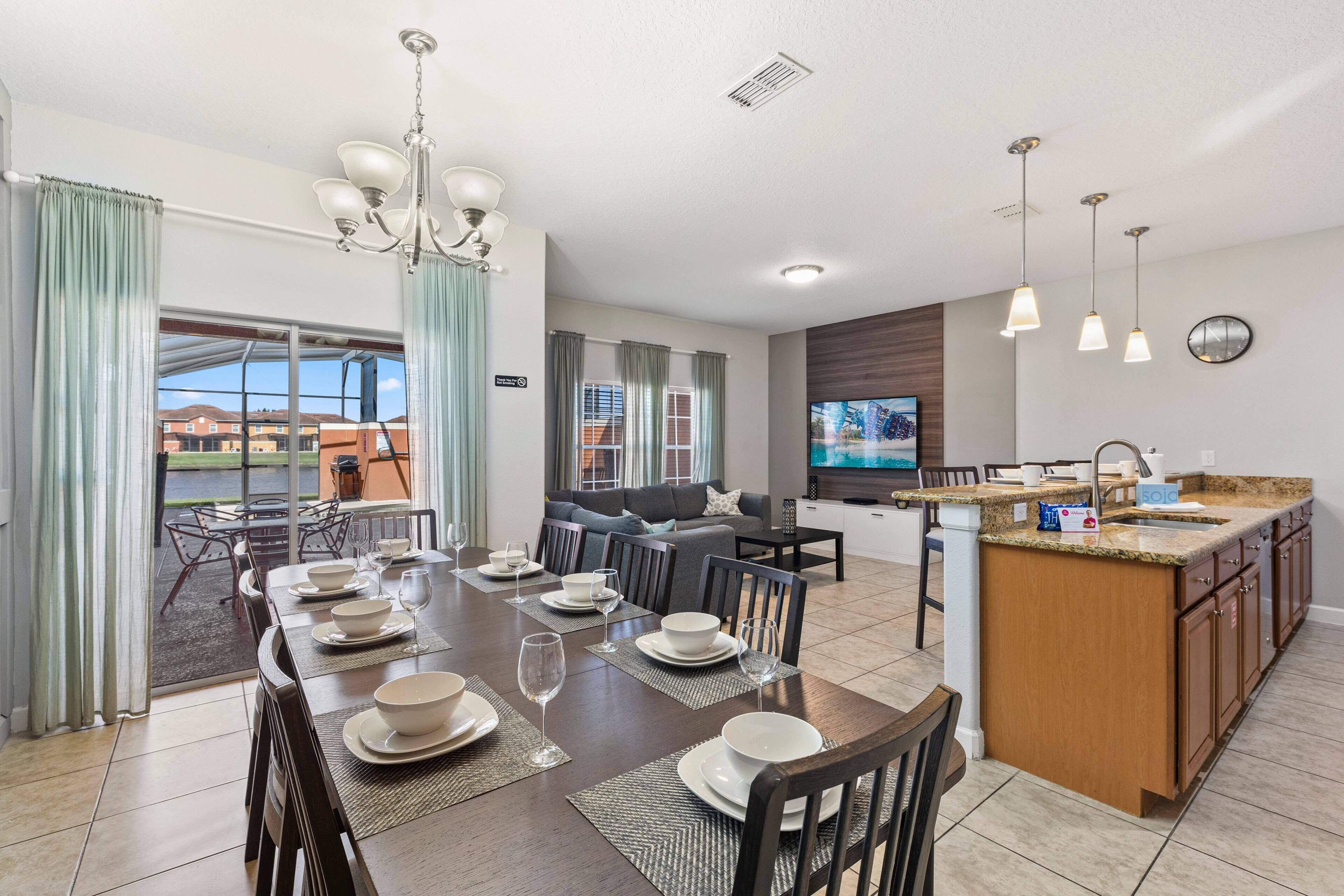 A modern kitchen with a central island, dining area with a set table, and a living room with a sofa and TV.