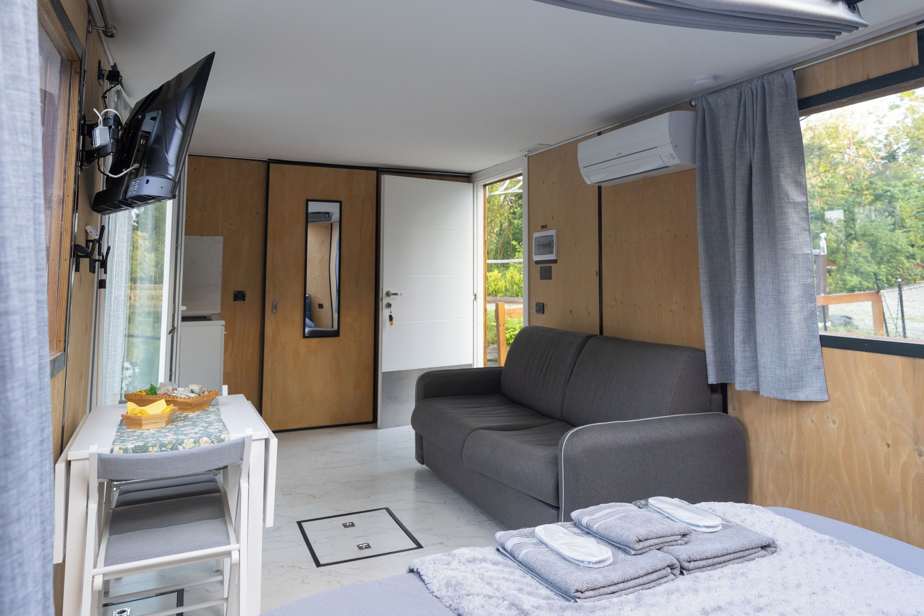 A modern room with a sofa, a small table with a basket, a bed with towels, a TV mounted on the wall, and a window with curtains.