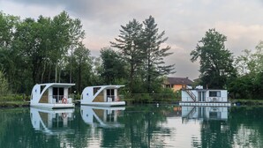Due houseboat con ponti e finestre, ormeggiate in acque calme, sullo sfondo alberi e una casa.