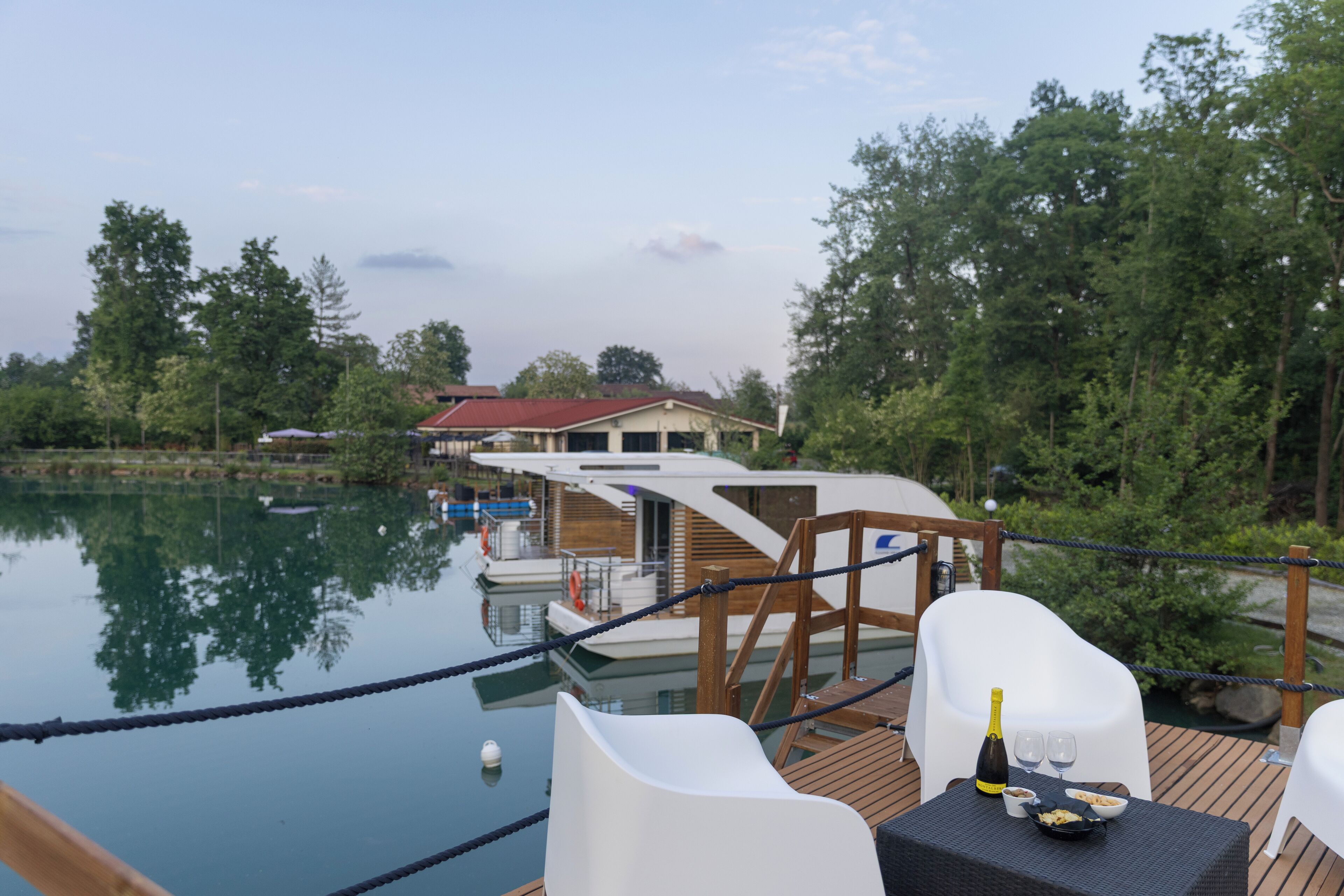 A deck with a table set for two, overlooking a calm lake with boats and a house in the background.