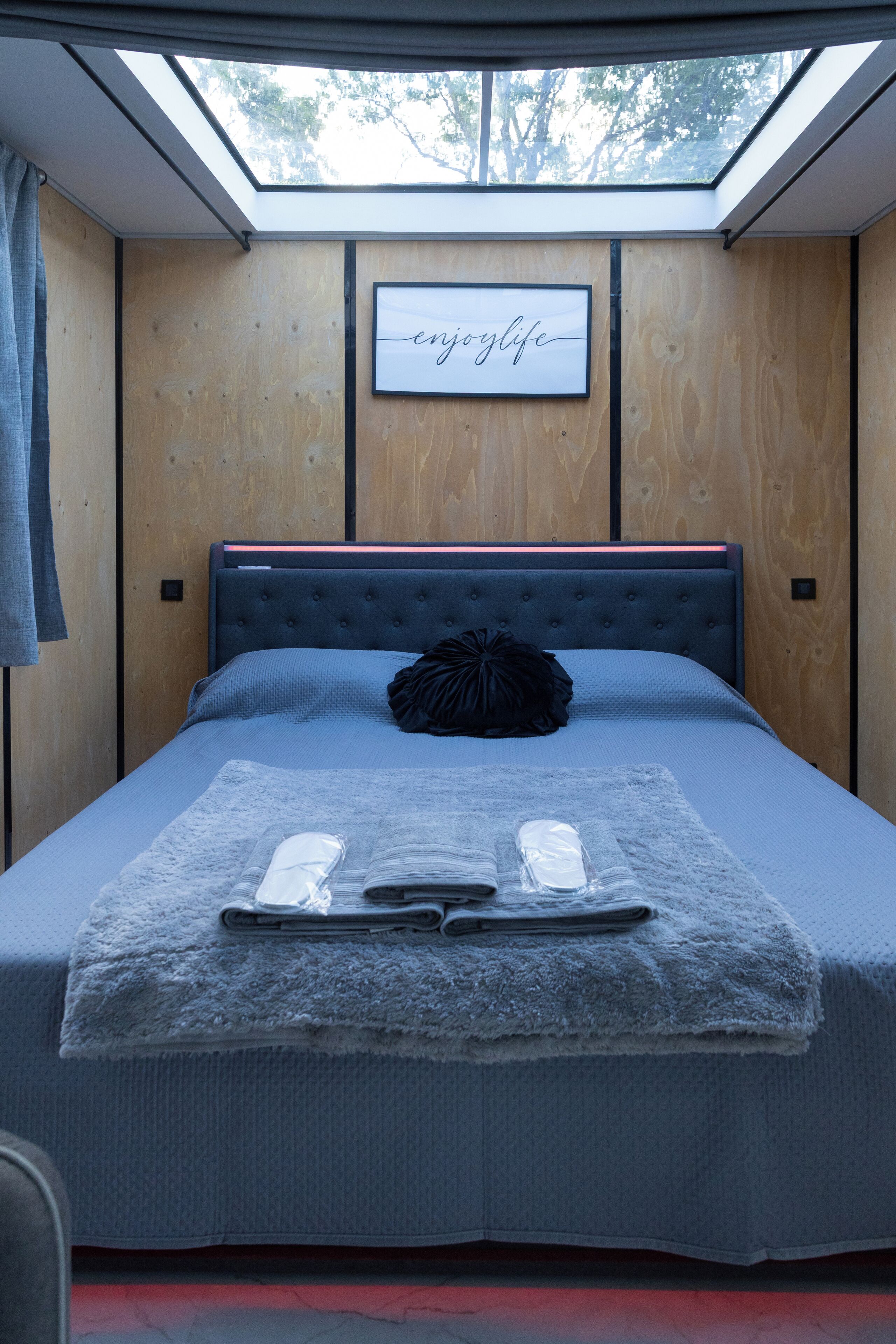 Une chambre avec un puits de lumière, un lit recouvert d’une courtepointe bleue, un oreiller noir et un cadre décoratif accroché au mur.