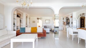 A spacious living area with a white sofa, a coffee table, and a dining table with chairs. There are bookshelves and a chandelier. - 4 bedroom house with pool, garden and beautiful views of the valley and the sea. (Santa Úrsula)