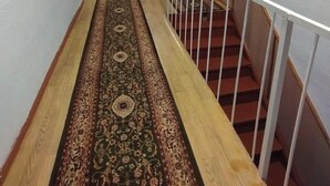 A narrow hallway with a wooden floor and a patterned carpet. A staircase with a white railing is on the right. - Jakesh (Karakol)