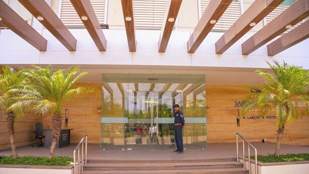 A hotel entrance with a security guard, palm trees, and a staircase leading up to the entrance. - Ambar Sarovar Portico (Gandhidham)