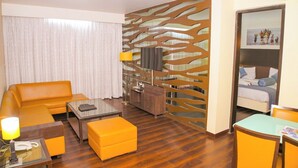 A hotel room with a sofa, ottoman, coffee table, TV, and a bed with a view. - Ambar Sarovar Portico (Gandhidham)