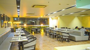 A modern dining area with long tables, cushioned seating, and a view through large windows. - Ambar Sarovar Portico (Gandhidham)