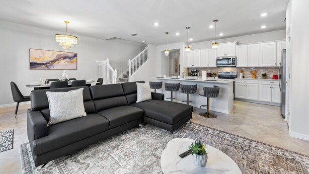 Ein modernes Wohnzimmer mit einer schwarzen Eckcouch, einem Essbereich mit weißem Tisch und Stühlen, einer Küche mit weißen Schränken und einer Kücheninsel sowie einer Treppe, die in einen weiteren Bereich führt.