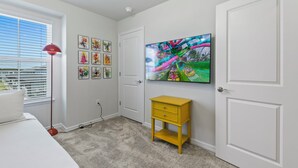 Ein Schlafzimmer mit einem Bett, einem Fernsehgerät an der Wand, einem kleinen gelben Nachttisch und einem Fenster mit Jalousien.