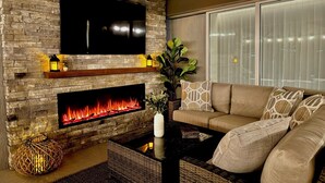 A living room with a stone fireplace, a flat-screen TV mounted above it, a sofa, and a large window with blinds.