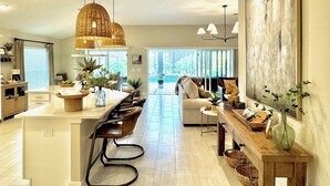 A modern kitchen with a central island, bar stools, pendant lighting, and a large window providing a view of the outdoors.