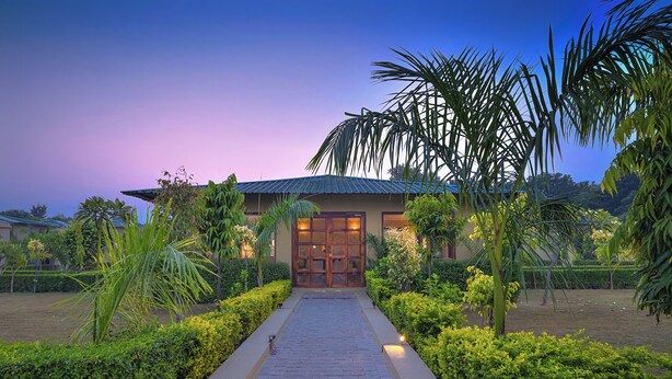 A house with a paved pathway leading to a wooden door, surrounded by lush greenery and palm trees. - Singhvilas Ranthambore-By Leisure Hotels (Sawai Madhopur)