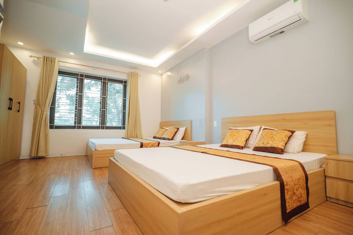 A hotel room with two beds, wooden flooring, a large window with curtains, an air conditioning unit, and a bedside table.