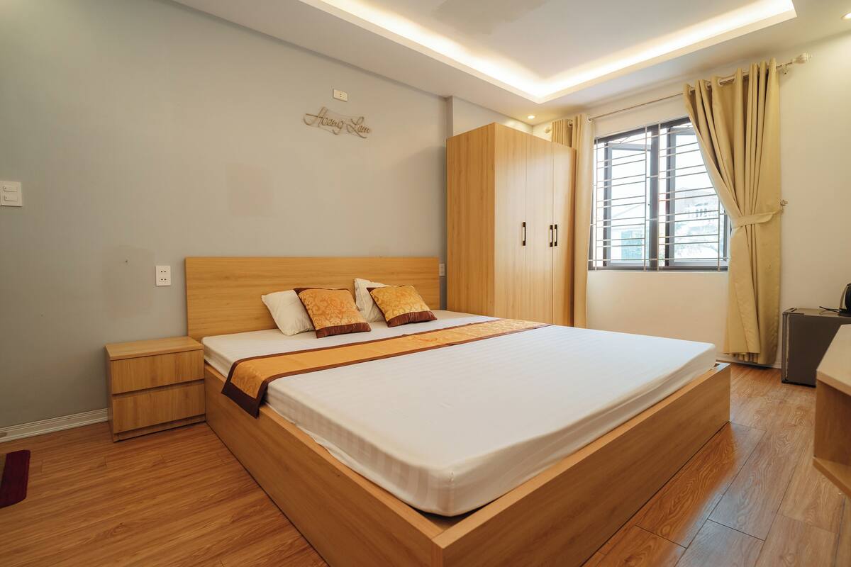 A modern bedroom with a large bed, wooden headboard, bedside tables, a wardrobe, and a window with curtains.