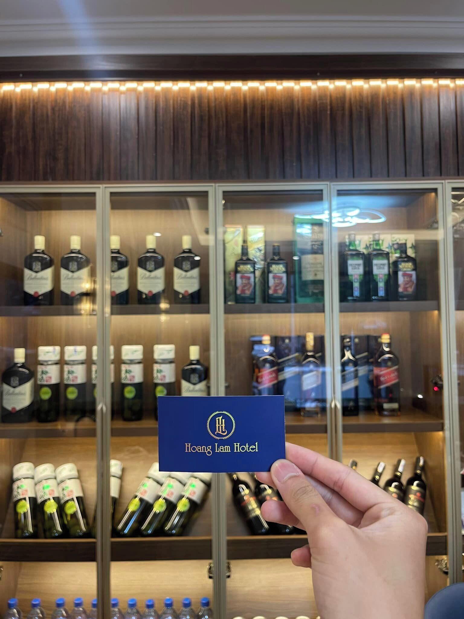 A hand holding a blue card with "Hoang Lam Hotel" written on it, in front of a display case filled with various liquor bottles.