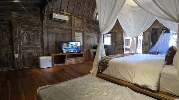 A bedroom with a wooden wall, a television, a bed with a canopy, and a small table. - Mara Inn (Nusa Dua)